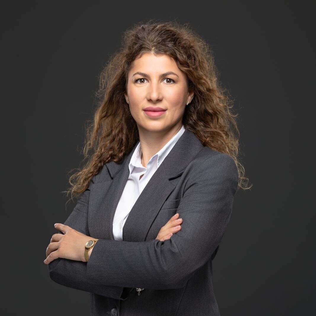 Felicia Lupu, the administrative support officer at JBV & Partners law firm in Luxembourg, standing with arms crossed in front of a grey background, ensuring efficient office operations.